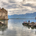 castle on a lake with mountains in the background and people on a pier